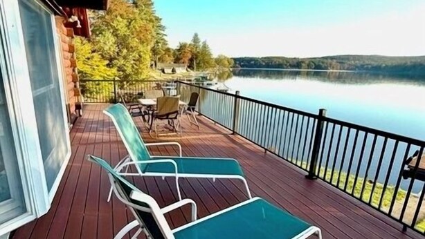 Una terraza de madera con dos sillones y una mesa, con vista a un lago tranquilo rodeado de árboles.