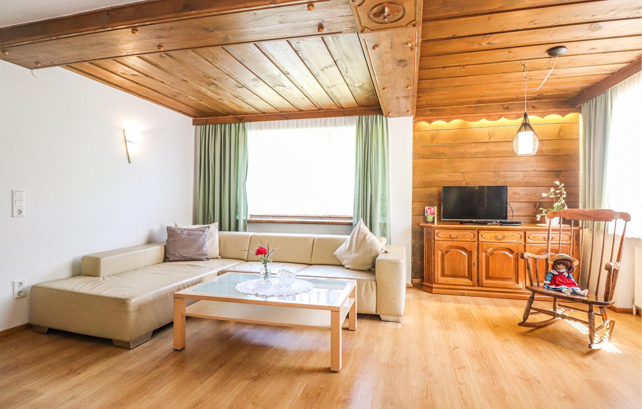 Uma sala de estar aconchegante com teto de madeira, um sofá, uma mesa de centro de vidro, uma televisão sobre um armário de madeira e uma cadeira de balanço.