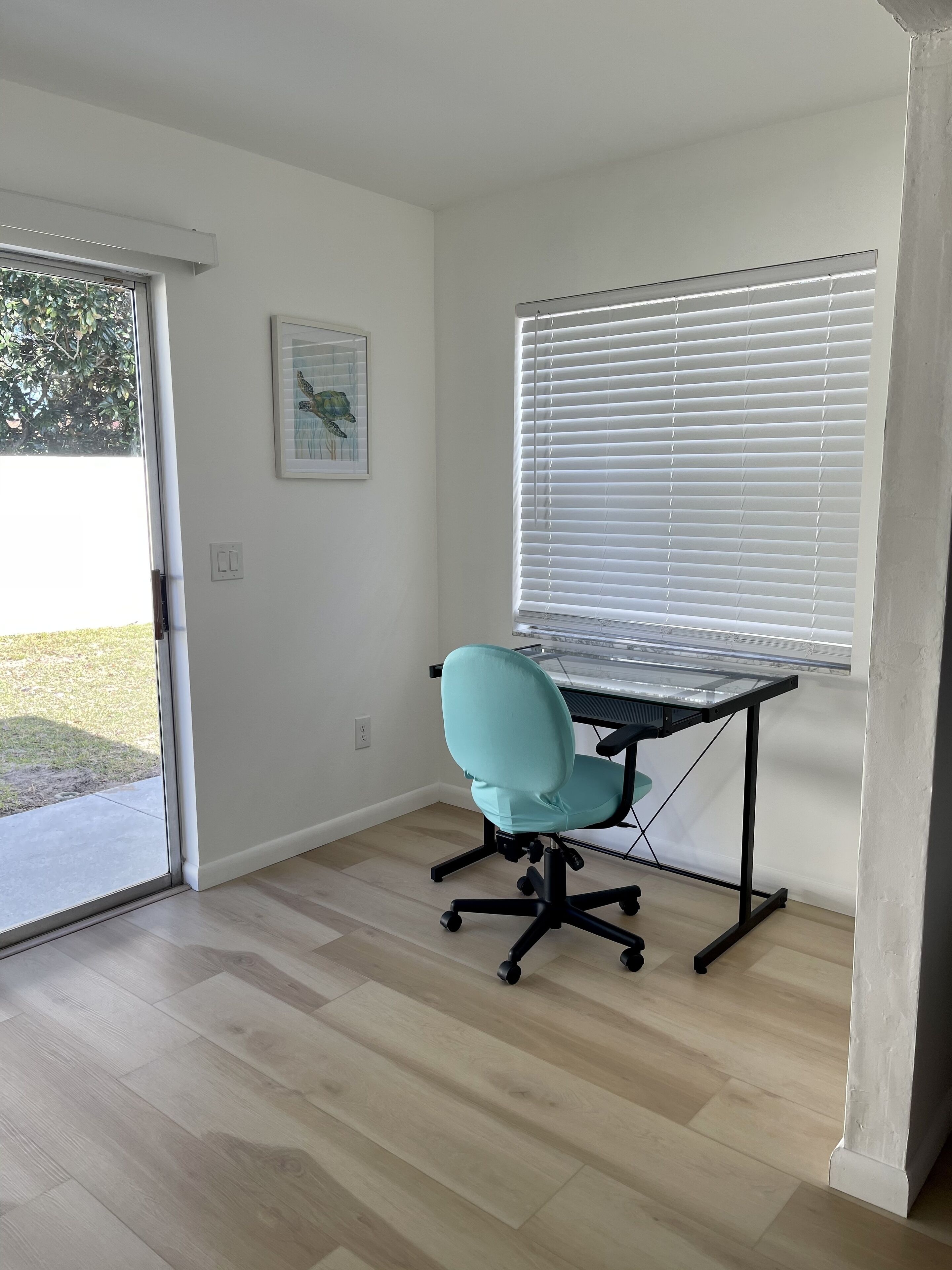 A room with a teal office chair, a glass-top desk, white walls, a window with blinds, and a door leading outside.