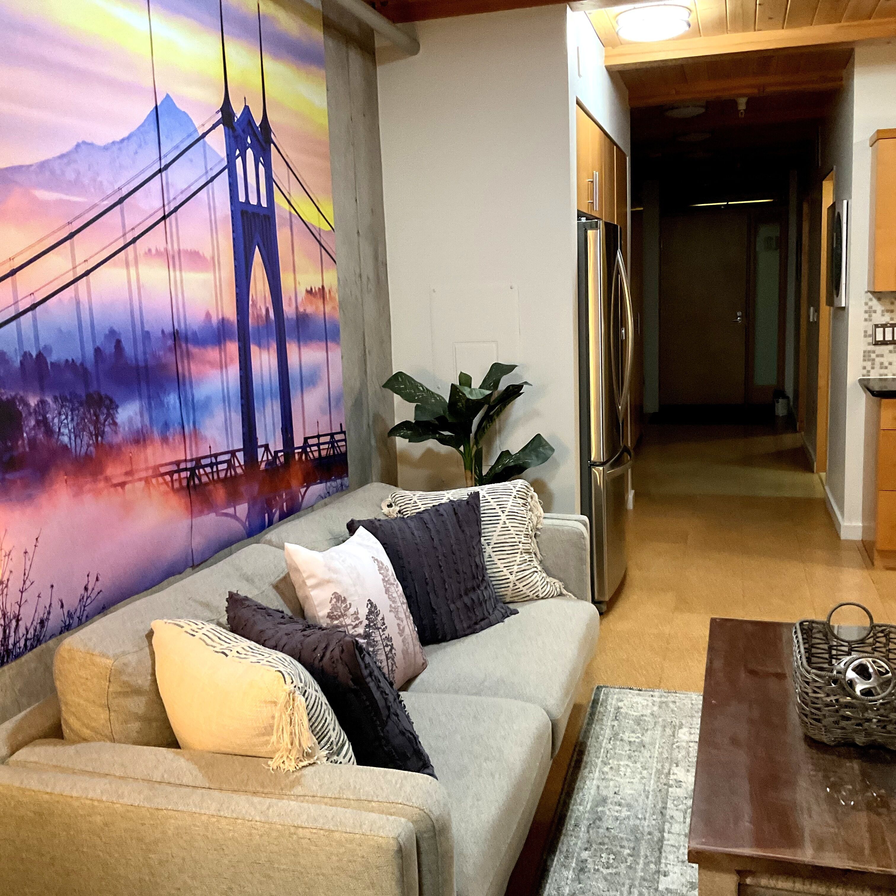 A living room with a sofa, a plant, and a painting of a bridge.