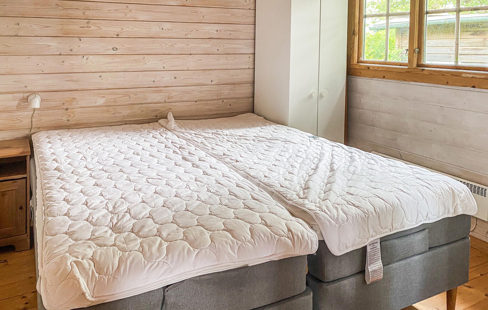 Un lit avec un matelas blanc matelassé, dans une pièce aux murs en bois et dotée d’une fenêtre.