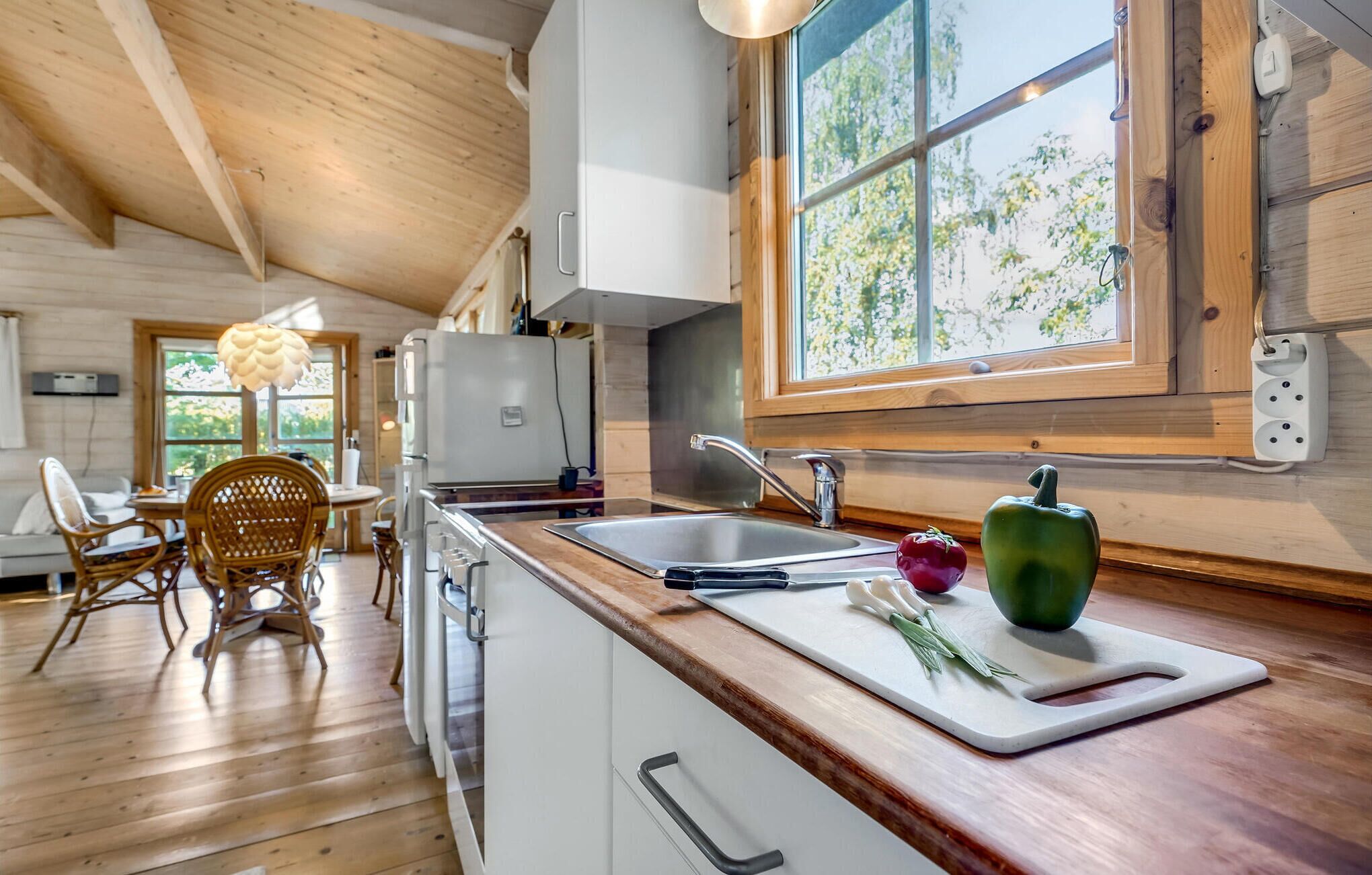 Une cuisine avec un plafond en bois, une grande fenêtre, un évier et un coin repas avec des chaises en osier.