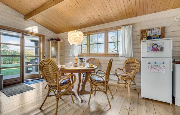 Une cuisine chaleureuse avec un plancher en bois, une table à manger entourée de chaises en osier, un réfrigérateur blanc et de grandes fenêtres avec des rideaux.