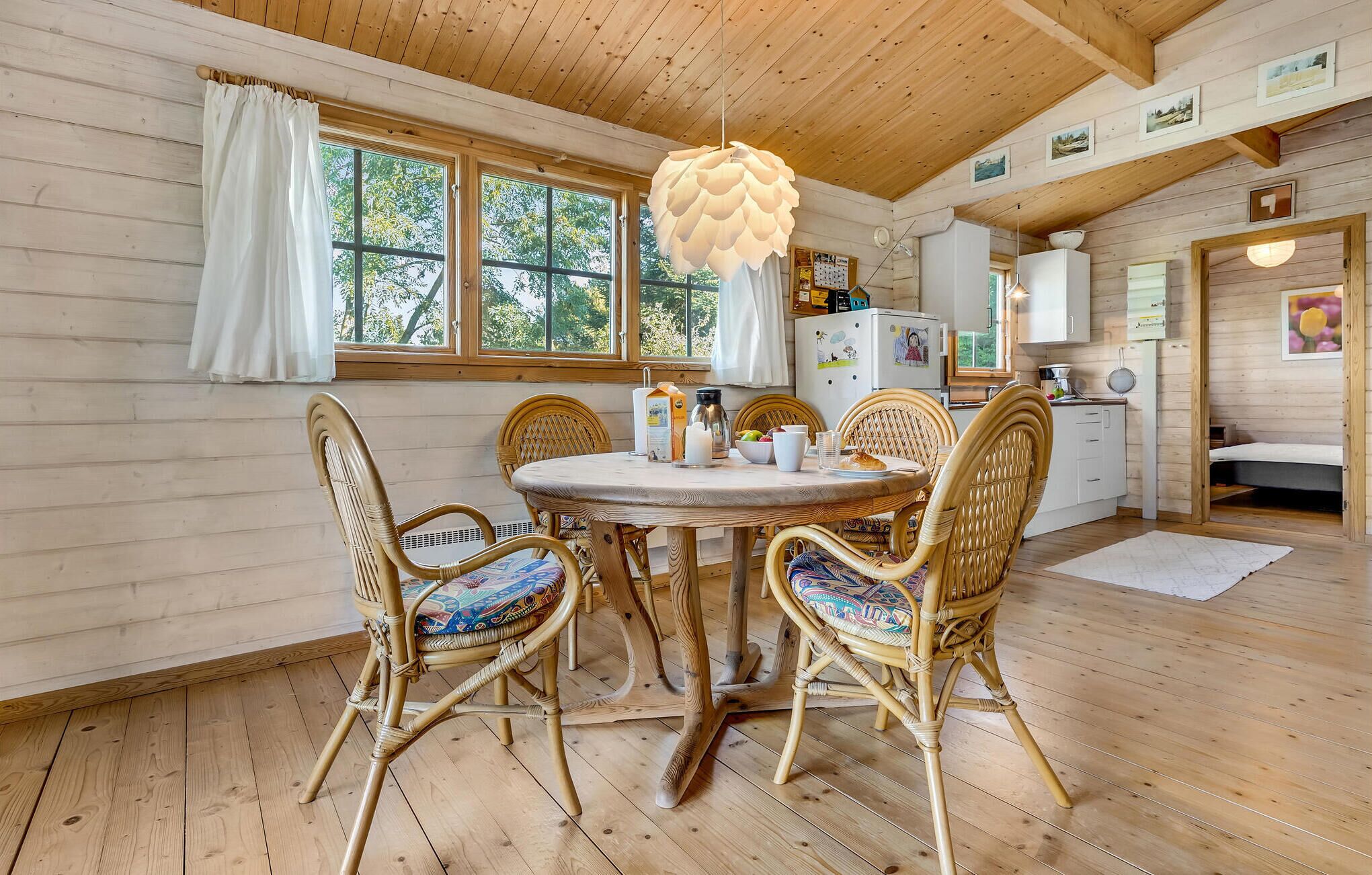 L’intérieur chaleureux d’un chalet en bois comprend un coin repas avec des chaises en osier et une table ronde, une cuisine avec des meubles blancs et de grandes fenêtres qui laissent entrer la lumière naturelle.
