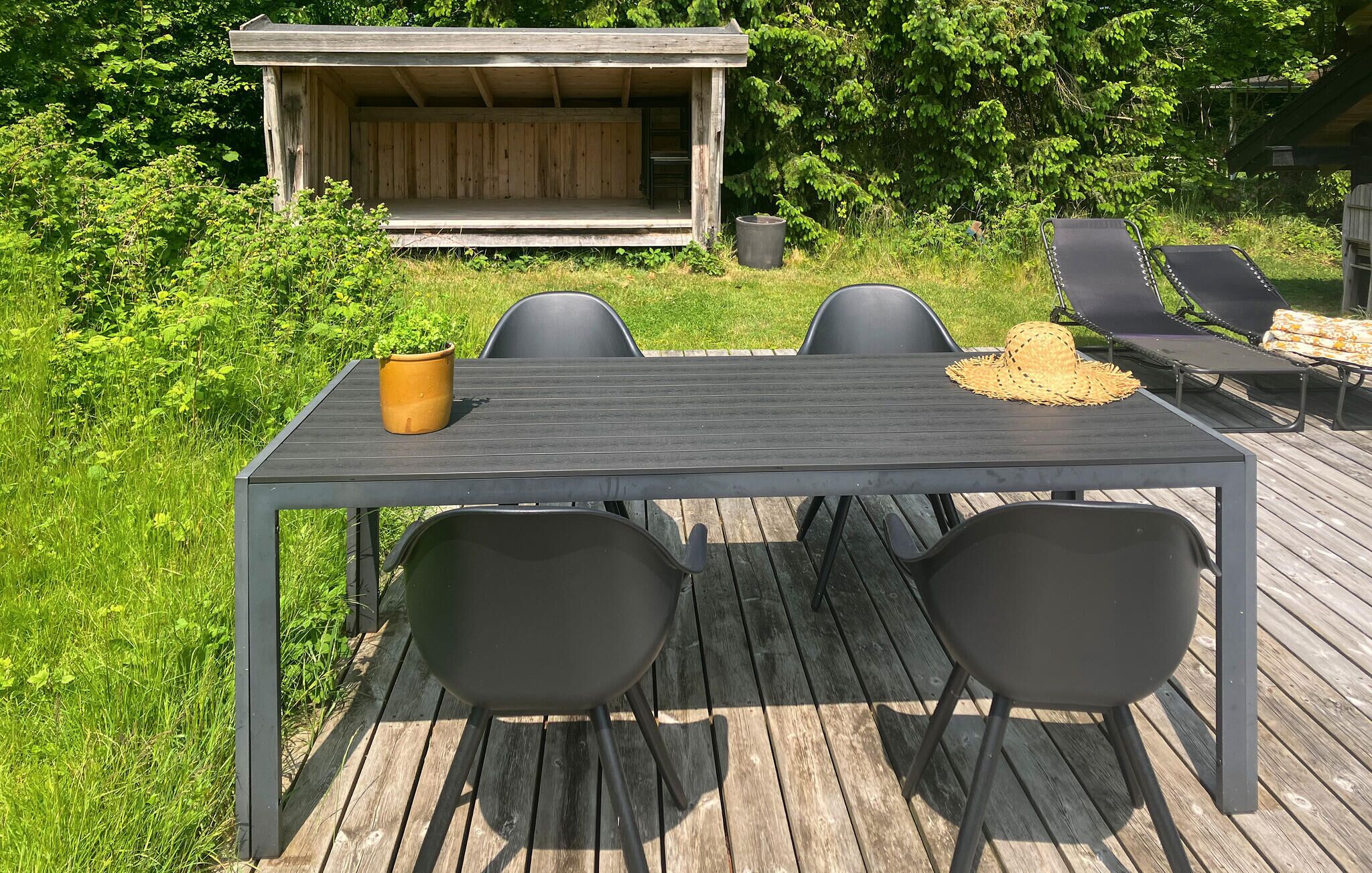 Una terraza de madera con una mesa y cuatro sillas, una pequeña construcción de madera al fondo y una planta en maceta sobre la mesa.