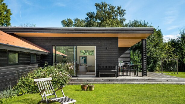 Een modern huis met een houten gevel, een glazen deuropening, een terras met een tafel en stoelen, en een gazon.