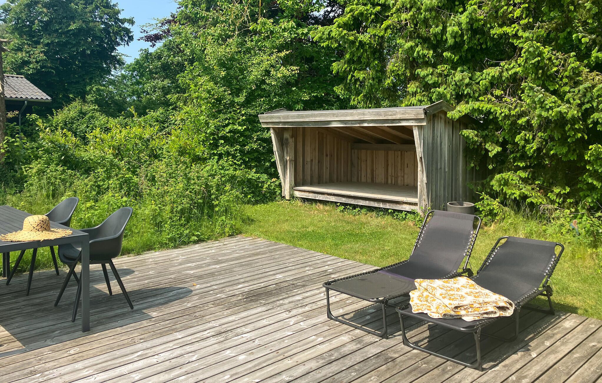 En træterrasse med et bord og stole, et overdækket siddeområde og frodigt grønt i baggrunden.