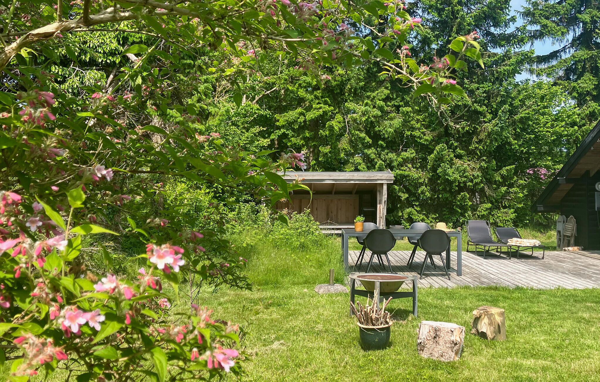 Une terrasse en bois avec une table et des chaises, entourée d’une végétation luxuriante et de plantes en fleurs.