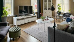 Una sala de estar moderna con una televisión de pantalla plana, una mesa de centro redonda, un sofá gris y un otoman tapizado.