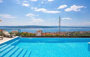 Ein Swimmingpool mit klarem, blauem Wasser, eine Lounge-Bereich mit Stühlen und einem Tisch sowie ein Blick auf eine Küstenstadt und Berge im Hintergrund.