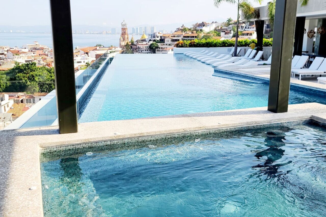 Una piscina infinita larga y estrecha con agua azul cristalina, rodeada de sillones de descanso y vistas a un paisaje urbano con una torre destacada.