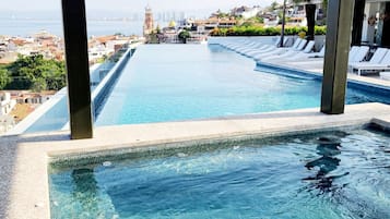 Una piscina infinita larga y estrecha con agua azul cristalina, rodeada de sillones de descanso y vistas a un paisaje urbano con una torre destacada.