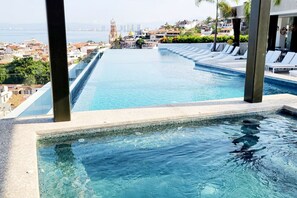 Una piscina infinita larga y estrecha con agua azul cristalina, rodeada de sillones de descanso y vistas a un paisaje urbano con una torre destacada.