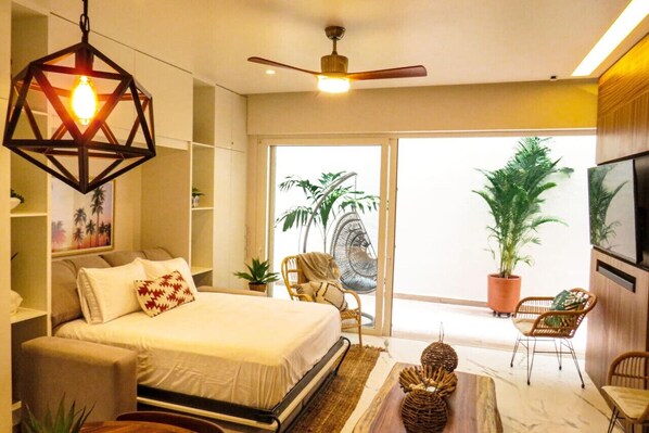 Un dormitorio moderno con cama, ventilador de techo, lámpara colgante, puerta corrediza de vidrio que da a un balcón con plantas y televisor montado en la pared.
