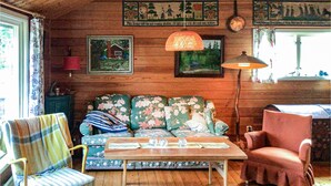 Un cuarto acogedor con paredes de madera, un sofá floral, un sillón a rayas, una mesa con copas de cristal y una lámpara colgante.