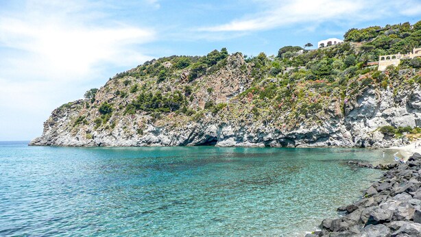 Paesaggio costiero con acque turchesi trasparenti, scogliere rocciose e un edificio sulla collina.
