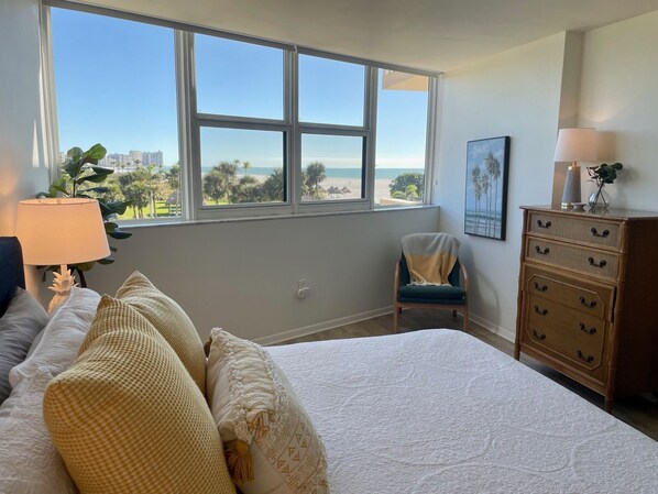 A bedroom with a large window offering a view of the beach and palm trees. There is a bed with white bedding and multiple pillows, a wooden dresser with drawers, and a chair with a blanket.
