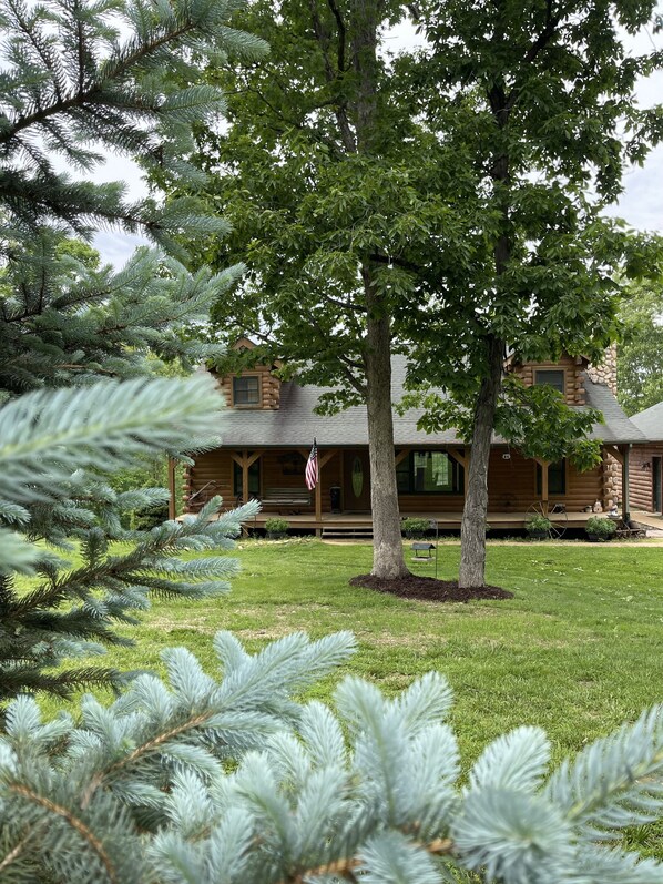 A log cabin with a porch, surrounded by a well-maintained lawn and evergreen trees. - Real Log Cabin with Seasonal Pool and Hot Tub with Meramec River Access (Bourbon)