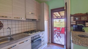 Et køkken med gaskogeplade, ovn og vask. Der er en dør, der fører ud til en balkon med en rød stol.