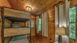 Quarto de cabana de madeira com beliches, uma mesa pequena com abajur e uma janela com cortinas.
