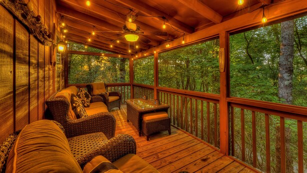 Um deck de madeira com móveis de vime e luzes de corda, com vista para uma área arborizada.