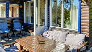 Een houten terras met een tafel, stoelen en een comfortabele bank, en grote ramen met blauwe omlijsting.