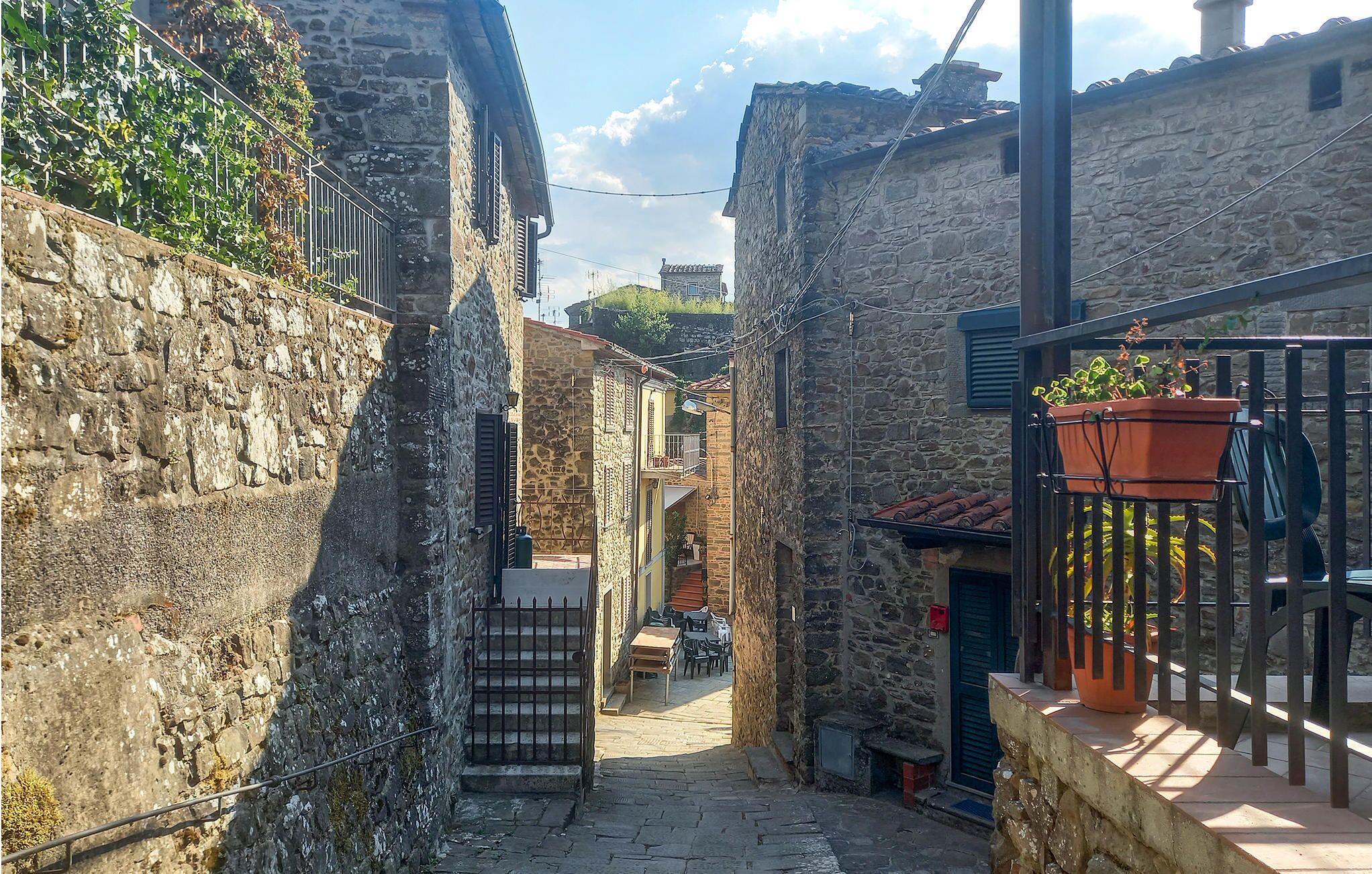 Ein schmaler Steinweg mit einer kleinen Außenbestuhlung und einer Treppe, die zu einem Hauseingang führt.