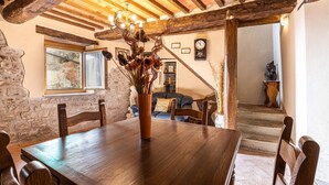 Ein rustikaler Essbereich mit Holztisch, Stühlen und einer Vase mit getrockneten Blumen. Eine Treppe führt in einen anderen Raum mit einer Couch und einer Wanduhr.