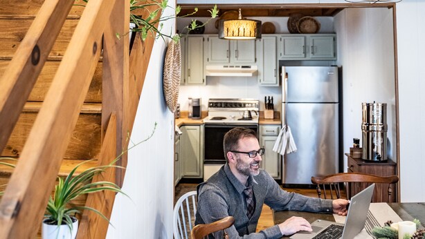 Un hombre usando una computadora portátil en una cocina rústica con vigas de madera, un comedor y una escalera.