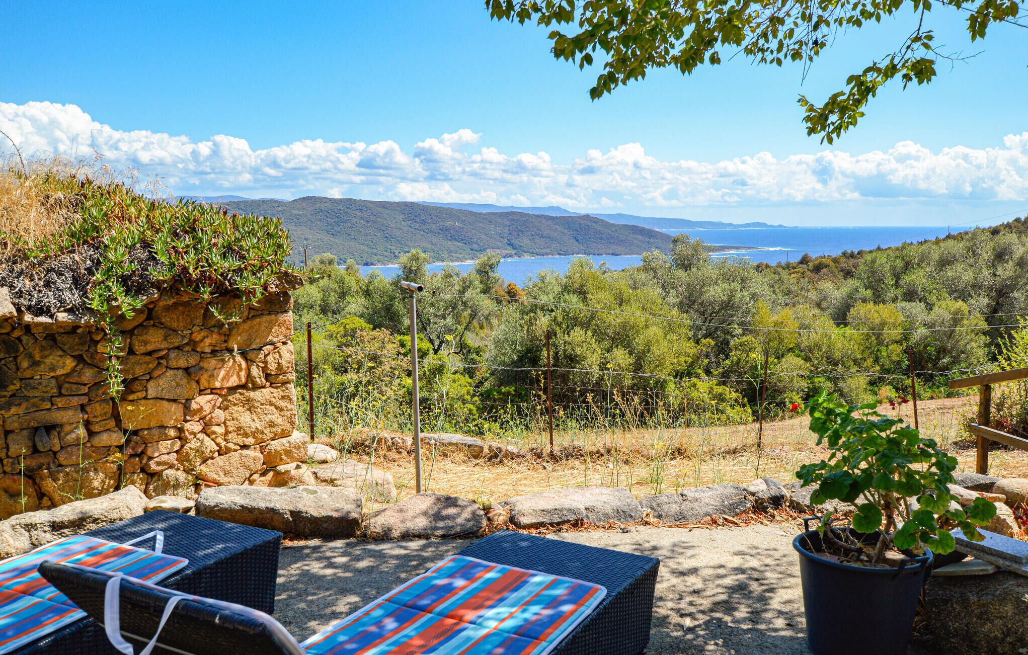 A stone wall with a potted plant, outdoor seating with a blue and red striped cover, and a view of a mountainous landscape with a body of water in the distance.