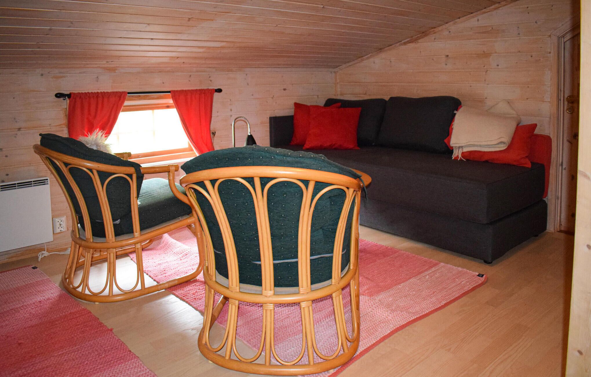 Une pièce chaleureuse avec un plafond en bois, un canapé agrémenté de coussins rouges et noirs, deux fauteuils en osier et un tapis rose.