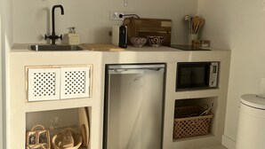 Een compacte keuken met een inbouwvaatwasser, een magnetron en een spoelbak. Boven de spoelbak zijn planken met servies en een klein mandje met keukengerei.