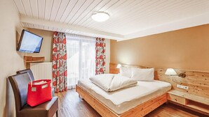 Ein Hotelzimmer mit einem hölzernen Bett, einem an der Wand befestigten Fernseher, einer roten Tasche auf einem Stuhl und einem Fenster mit Vorhängen.