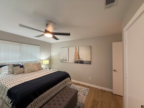 Une chambre à coucher comprenant un lit, un ventilateur de plafond, une fenêtre avec des stores, une lampe et une œuvre d’art représentant des voiliers, accrochée au mur.