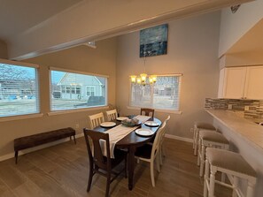 Un coin repas avec une table en bois, des chaises blanches et un banc. On y trouve des fenêtres avec des stores, un lustre et un tableau au mur.