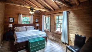 Un dormitorio de cabaña de madera con una cama, dos ventanas, un ventilador de techo y un baúl verde.