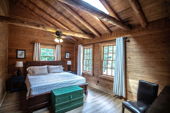 A wooden cabin bedroom with a bed, two windows, a ceiling fan, and a green trunk.