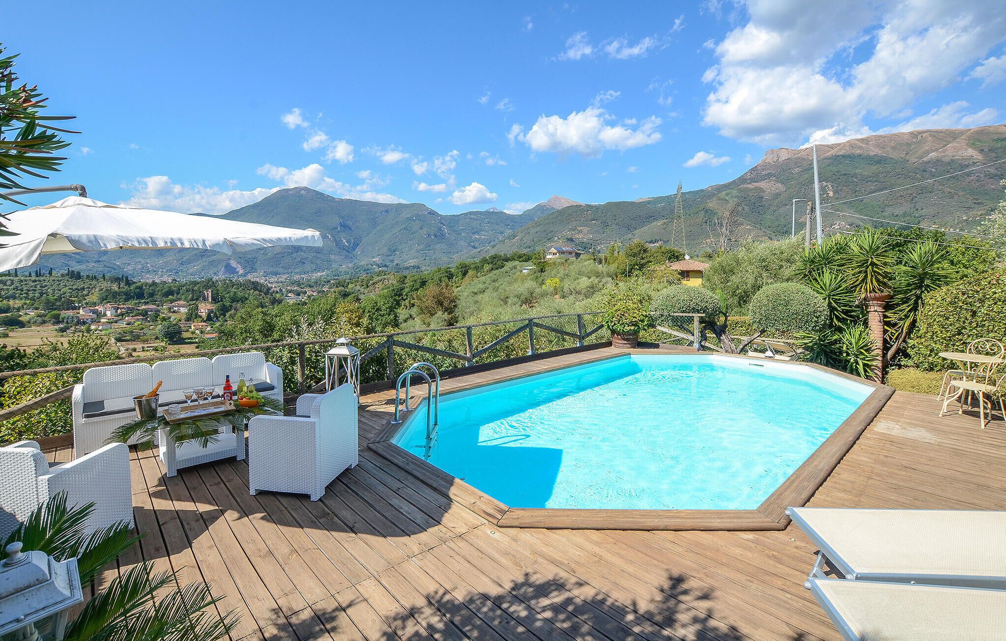 A pool area with a wooden deck, white lounge chairs, and a view of a village and mountains in the background.