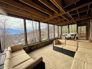A room with a stone wall, large windows, and a view of mountains. There are two beige sofas and a dining table with chairs.