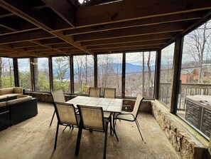 Ein überdachter Essbereich im Freien mit Tisch und Stühlen, große Fenster mit Blick auf Bäume und Berge.