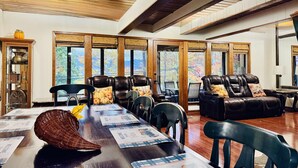 A spacious room with leather sofas, a long wooden table, and large windows offering a view of trees.