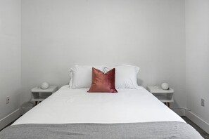 Une chambre à coucher au style minimaliste, avec un grand lit, du linge de lit blanc et un seul coussin décoratif.