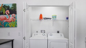 A laundry room with two white front-loading washers and dryers, a shelf with cleaning supplies, and a painting of flamingos on the wall. - 8 bedrooms - Stunner in Miami Design District! (Miami)