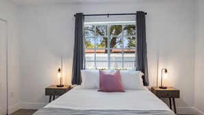 A bedroom with a large bed, two bedside tables with lamps, a window with curtains, and a view of trees and a fence outside. - 8 bedrooms - Stunner in Miami Design District! (Miami)