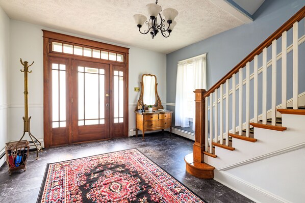 A spacious foyer with a large wooden door, a staircase with wooden railings, a chandelier, and a patterned rug.