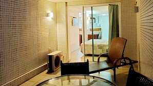 Une petite pièce carrelée, avec une table et des chaises en verre, un haut-parleur et une vue sur une chambre à coucher avec un lit et une fenêtre.