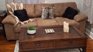 En stue med en brun sofa, to sorte og leopardmønstrede puder, et maleri af en hvid blomst på væggen, et træsofa bord med stearinlys, og en lille urtepotte.