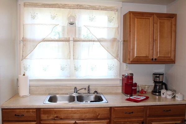 A kitchen with a double sink, wooden cabinets, a coffee maker, and a window with sheer curtains.