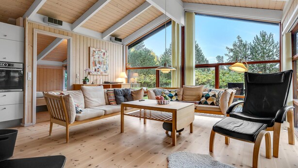 Amplia sala de estar con techo alto, vigas de madera, un ventanal con vistas a árboles, un conjunto de sofás, una mesa de comedor y una butaca de cuero negro.