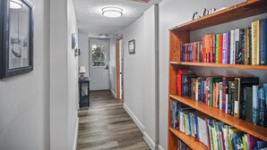 Un couloir avec une bibliothèque en bois remplie de livres, un tableau encadré au mur et une porte au fond.